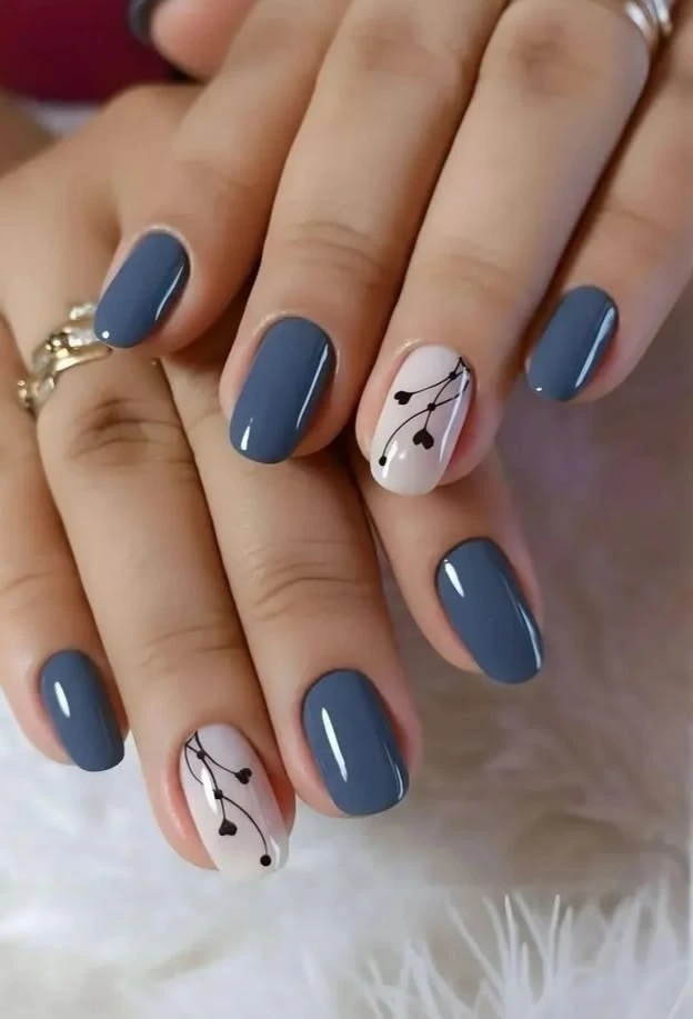 A close-up beauty shot of short nail ideas featuring a glossy slate blue manicure on short squoval nails with soft neutral accent nails decorated with black botanical vine art.