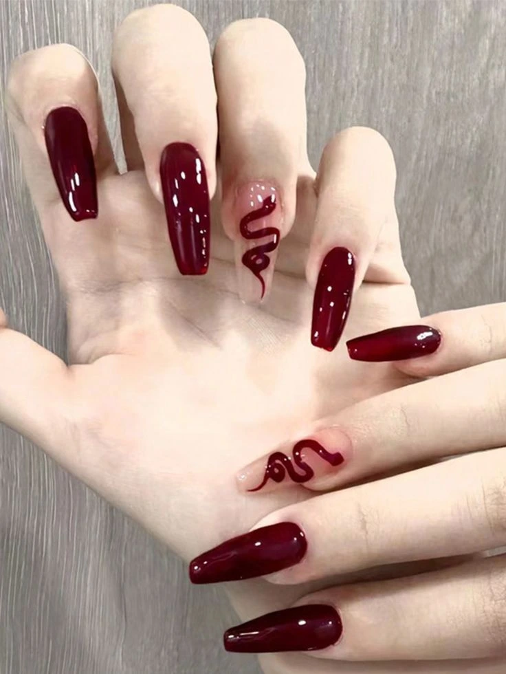 A close-up view of dramatic coffin-shaped nails featuring deep burgundy shades, nude accent nails, and sculpted glossy serpent details creating a luxurious gothic-inspired nail design.